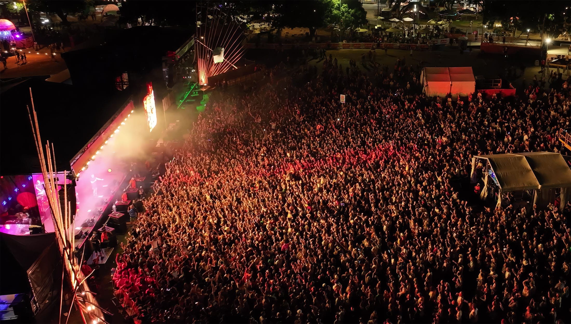 Aerial view of a crowded outdoor concert at night, with colorful stage lights illuminating the enthusiastic audience.